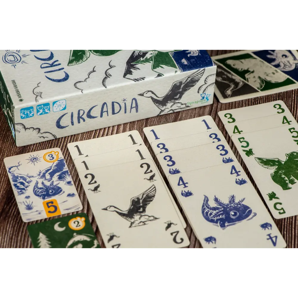 A boxed card game called Circadia with several cards and a small token laid out on a wood surface. The box and cards show blue and green illustrated graphics on an off-white background.