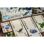 A boxed card game called Circadia with several cards and a small token laid out on a wood surface. The box and cards show blue and green illustrated graphics on an off-white background.