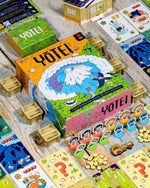 A colorful board game set labeled YOTEI arranged on a rustic wooden table, with the main illustrated box in the center and matching game components like cards, coins, tokens, and small furniture pieces spread around it.